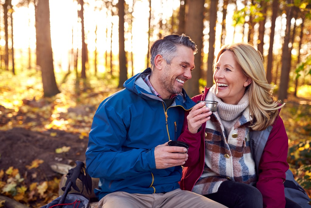 An older couple enjoys coffee from a thermos during a hike through a forest in fall.