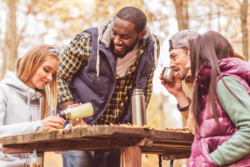 Group of smiling friends sitting at wooden table and pouring hot drink in autumn forest