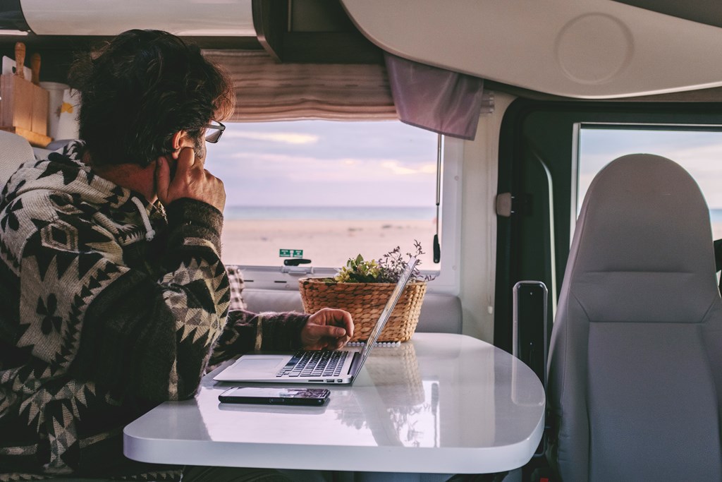 Man working inside camper van sitting at the table and looking outside the window the beautiful beach and ocean in background. 