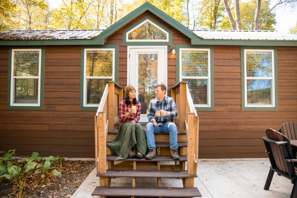 Two people sitting on the steps of a Deluxe Cabin holding coffee cups in the fall at Coshocton KOA Holiday, Ohio