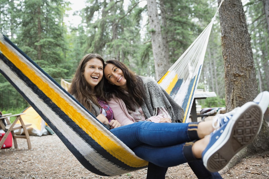 Cheerful mother and daughter sitting on hammock at campsite.