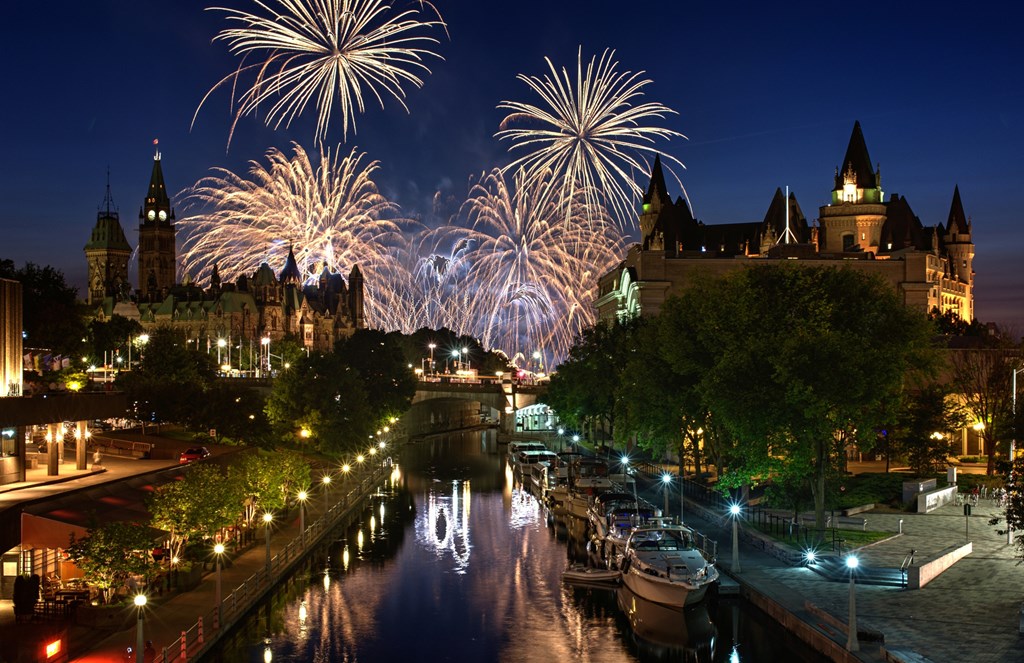 A colour image featuring a summer fireworks display festival in downtown Ottawa Canada. In the foreground are the Parliament Buildings, Rideau Canal a UNSECO world heritage site, and the Chateau Laurier hotel.