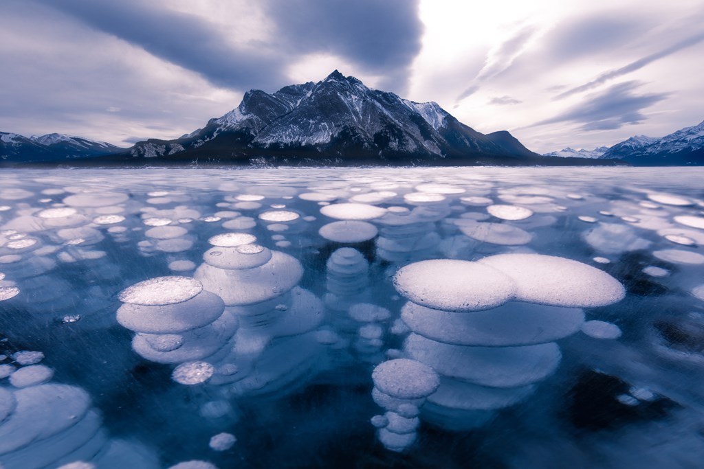 Layered ice bubble in frozen Lake Abraham with mountains and cloudy sky in the background.