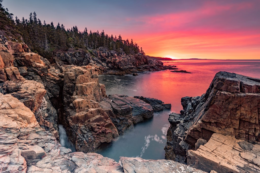 A landscape scene with a pink sunset over the ocean and rocky coastline in Acadia National Park in Maine