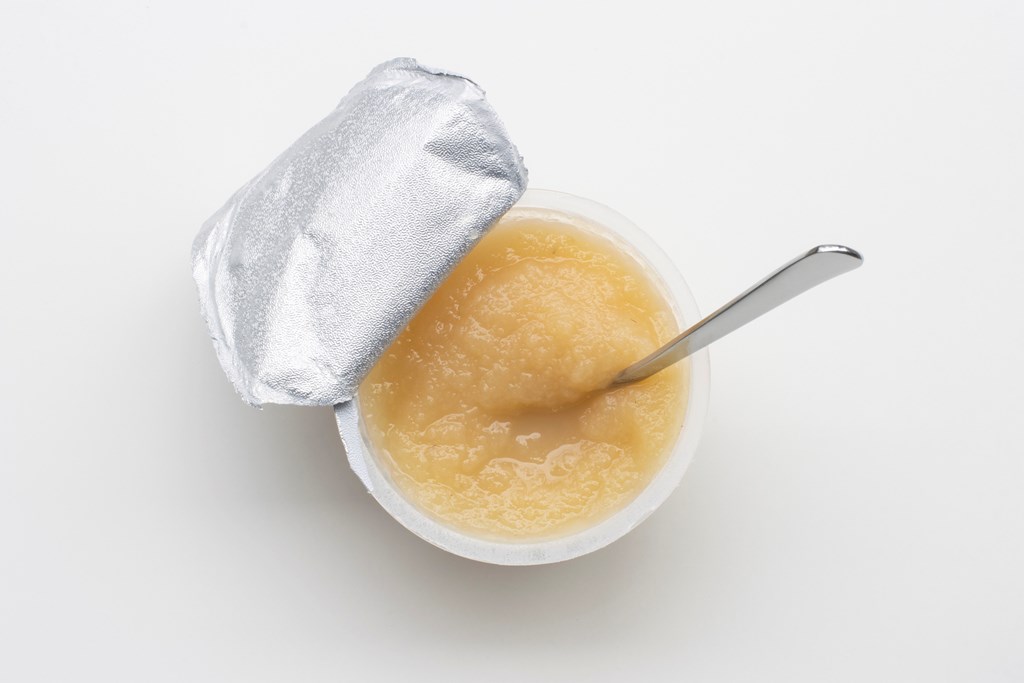 Top view of an opened cup of unsweetened applesauce with a spoon in it, isolated on a white background.