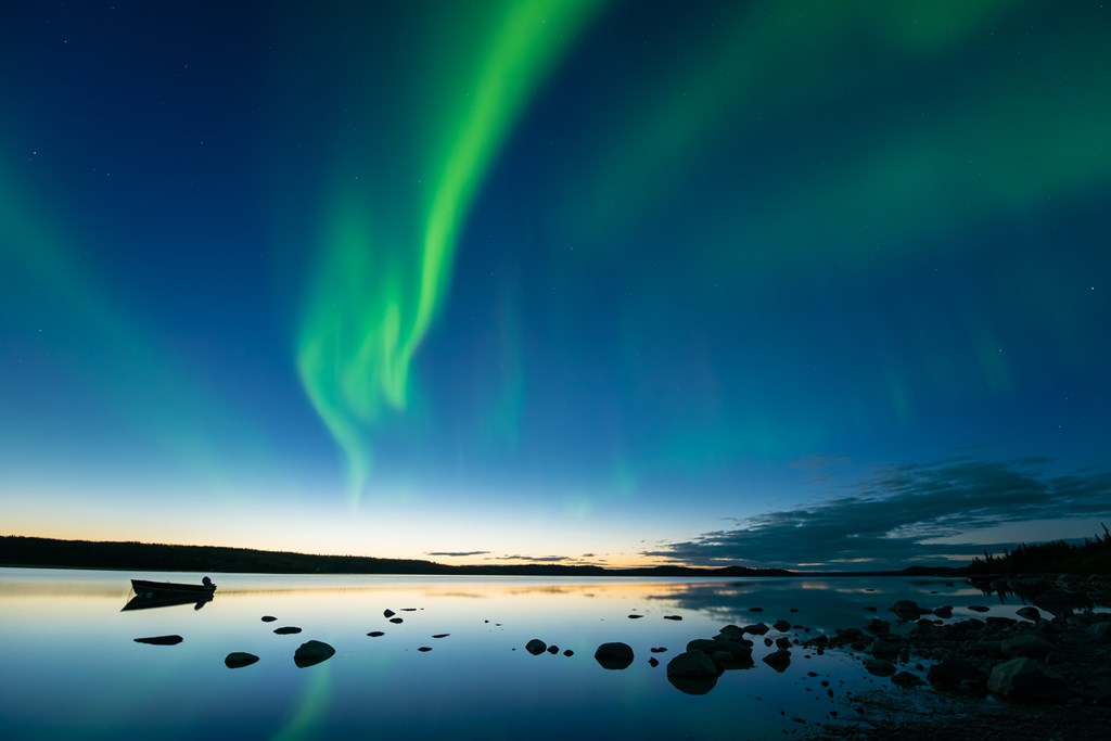 Bands of curvy aurora borealis appear over a northern rocky lake right after sunset.