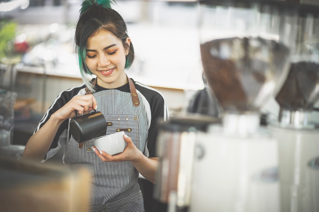Young female barista finishing coffee preparation by pouring foam in a mug.