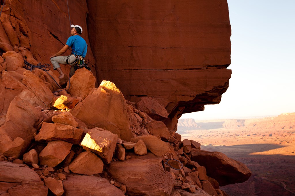 A young man sitting on a rock belays his partner while rock climbing in Utah.