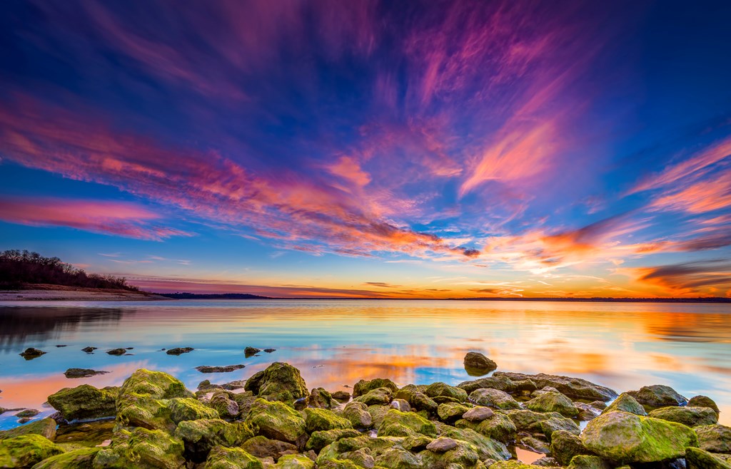 Amazing multicolored sunset over Benbrook Lake in Fort worth, TX featuring vivid green mossy rocks in the foreground.