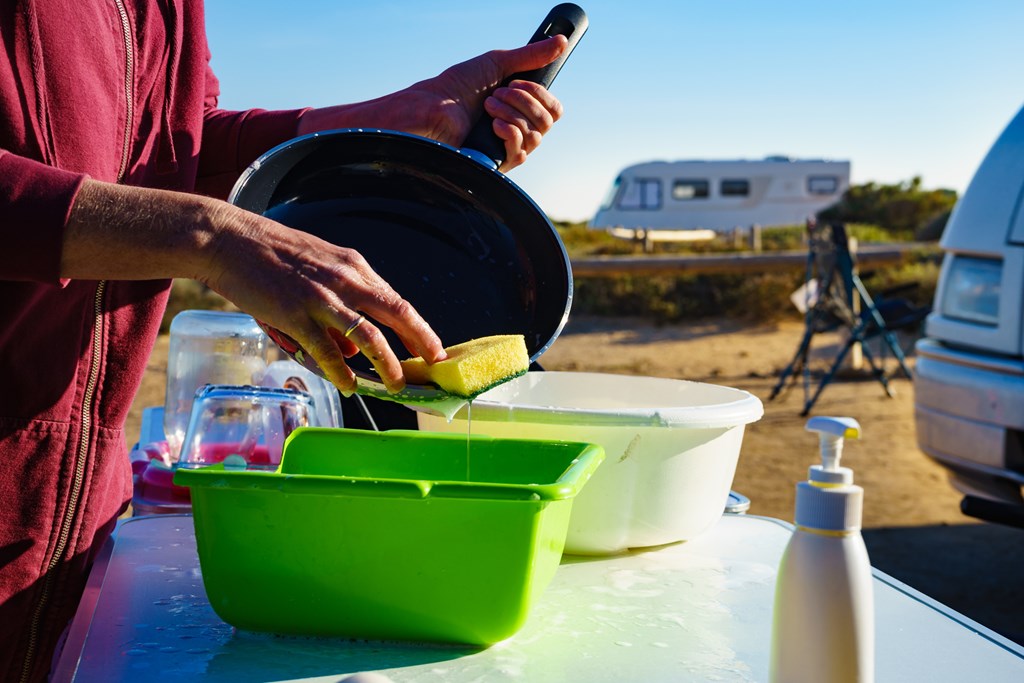 Mature woman washing up dishes in bowl on fresh air. Dishwashing outdoor on camping site, sea shore