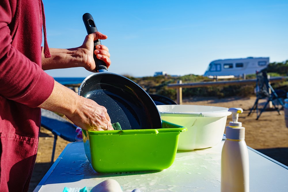 Mature woman washing up dishes in bowl on fresh air. Dishwashing outdoor on camping site, sea shore
