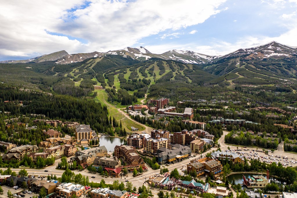 A drone photo of Breckenridge, Colorado in summer shows a quaint city surrounded by deep green mountains. On one mountain, you can see green outlines of ski trails.