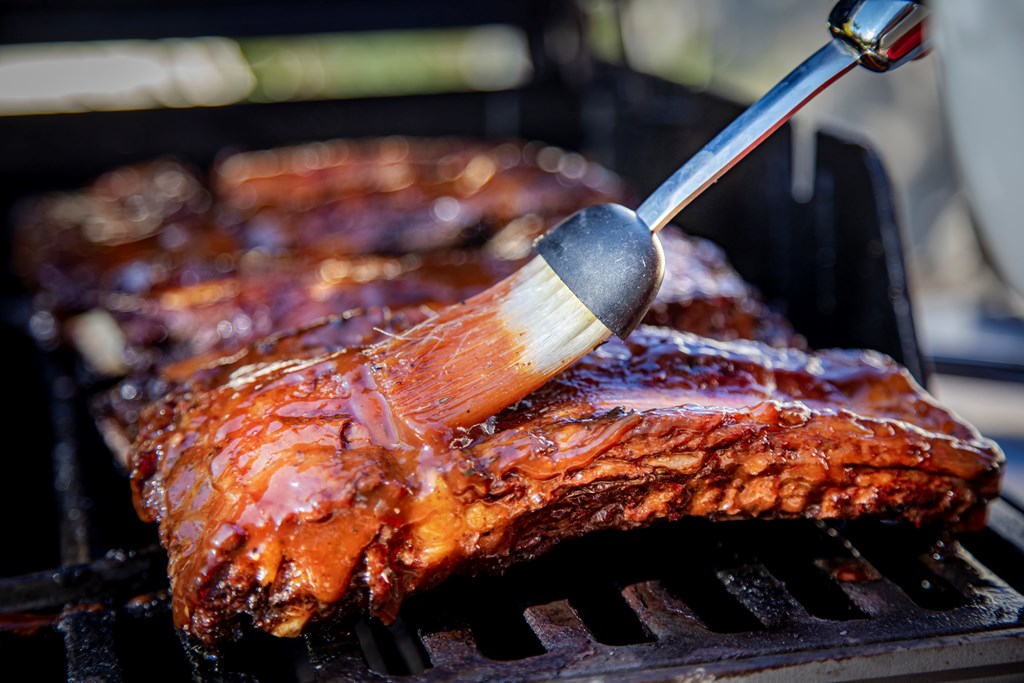Beef ribs being basted with bbq sauce on a grill with a basting brush.