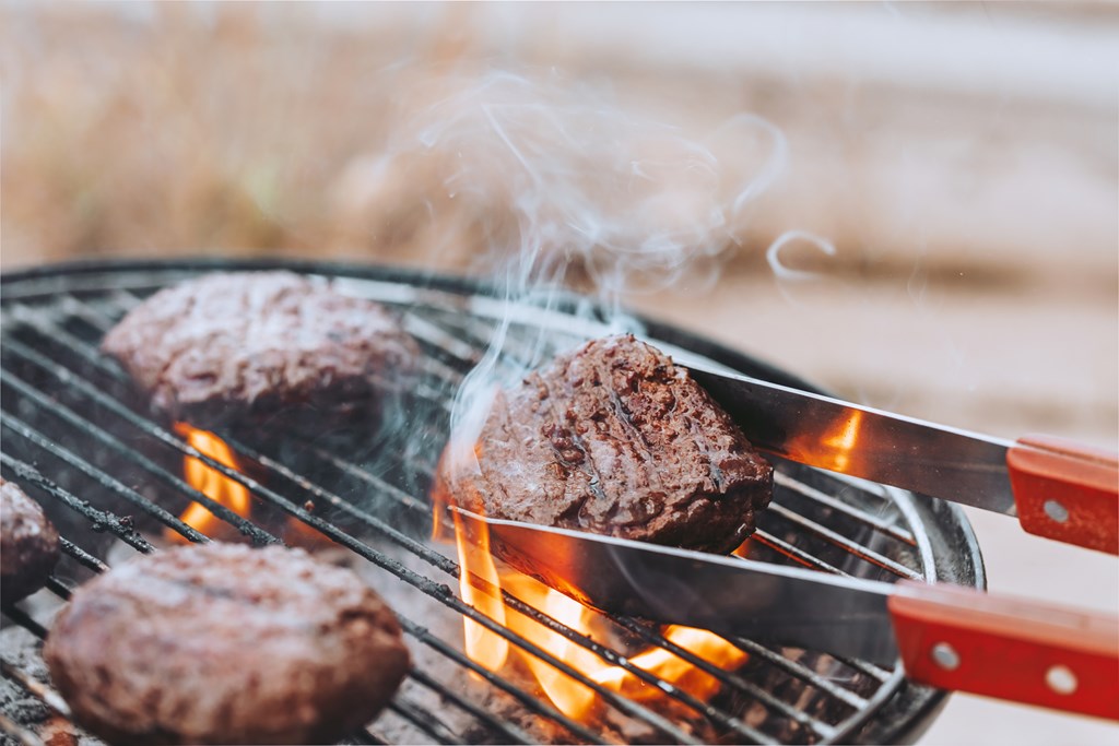 Closeup of tongs flipping a burger on a charcoal grill with flames. 