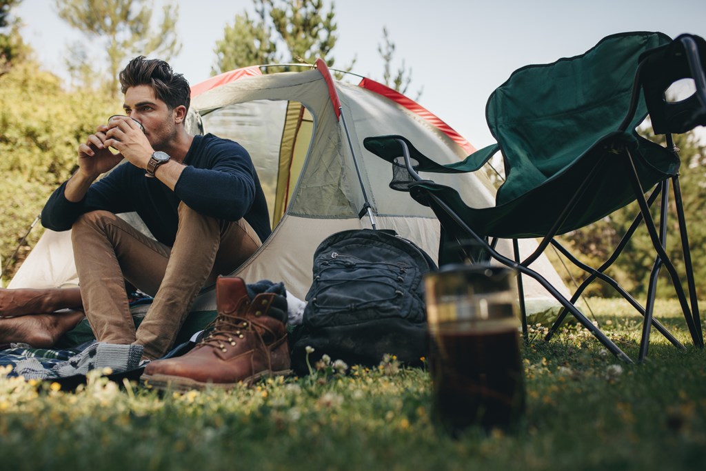 Young man sitting in his tent at campsite and drinking coffee. 