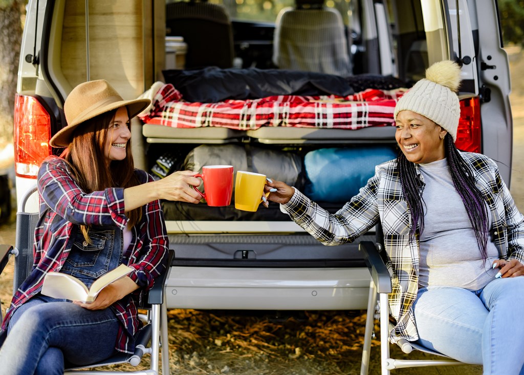Two female travelers clinking mug of hot drinks while sitting in chairs near camper in forest and enjoying autumn weekend.
