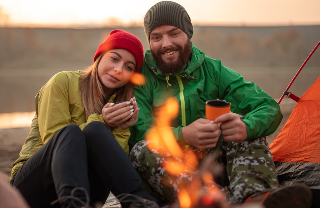 Young man and woman with cups of hot beverage relaxing near the campfire.