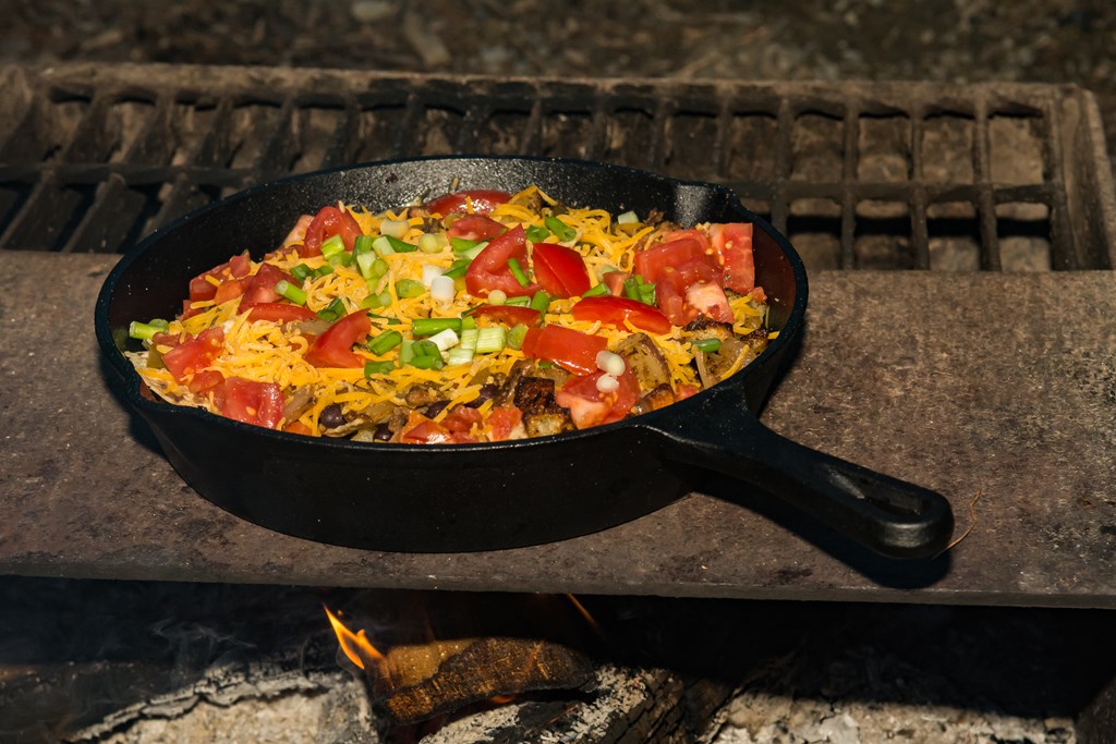 A cast iron skillet of nachos with cheese, tomatoes and ground beef sits over a campfire.