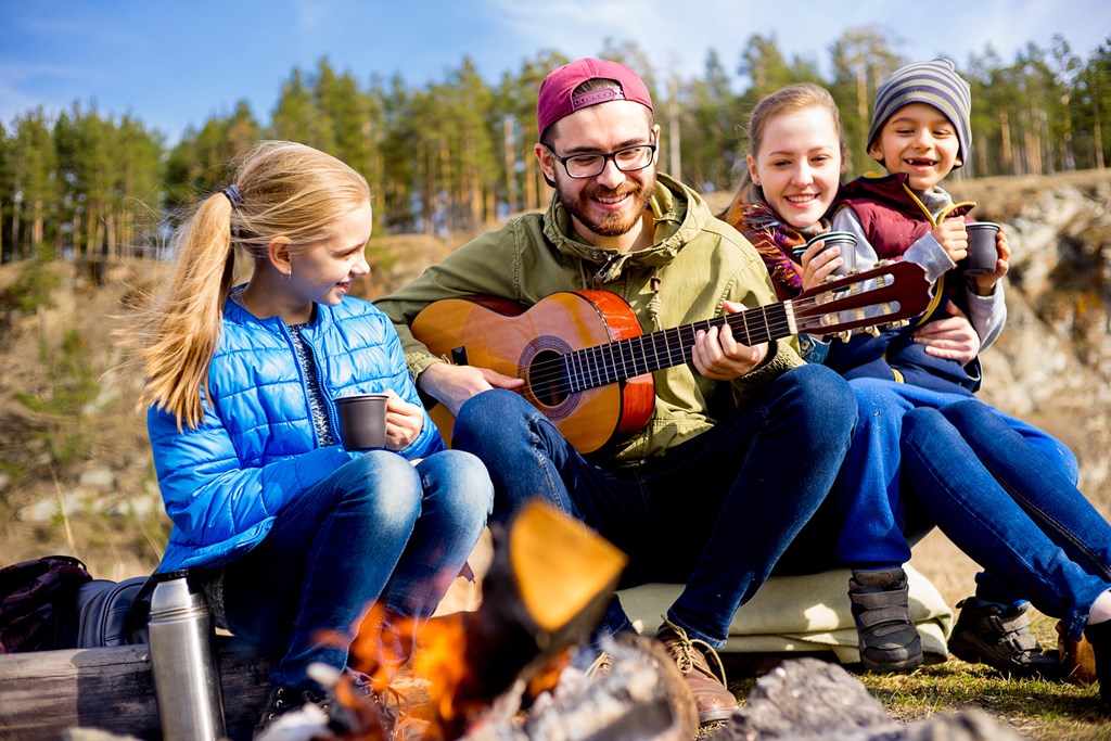 Happy family of four sits by a campfire while dad plays the guitar and sings campfire songs.