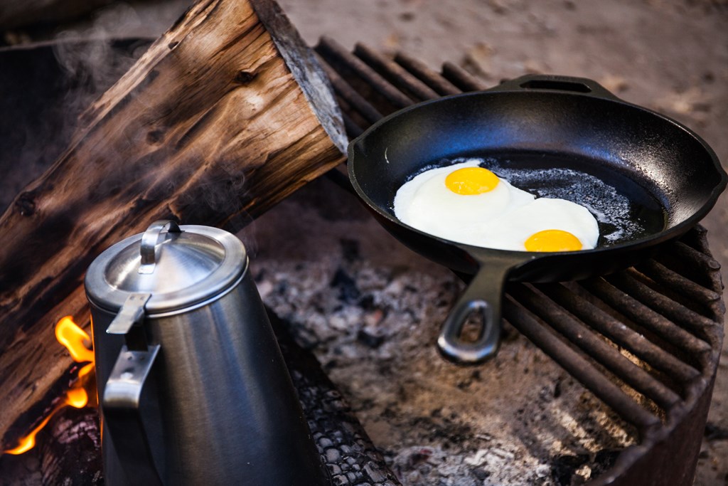 Two eggs in a cast iron frying pan and a coffee pot over a campfire.