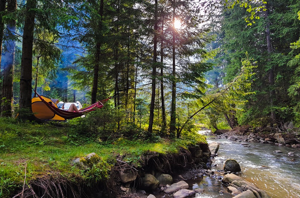Woman resting on a hammock at a green forest campsite by a mountain stream.