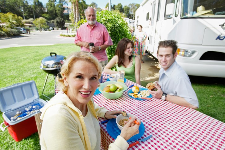 Senior Couple Sit Outdoors at a Table in Front of Their Motor Home, Having Breakfast With Their Teenage Grandchildren