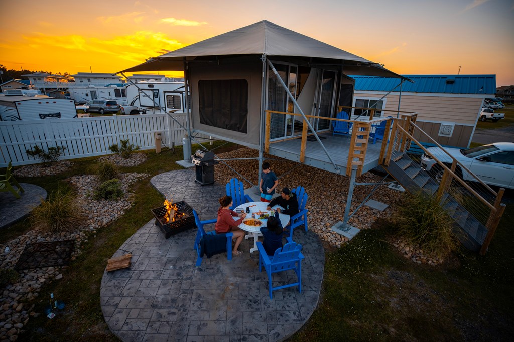 A family sits down to dinner on an upscale patio in front of an elevate glamping tent at a KOA campground.