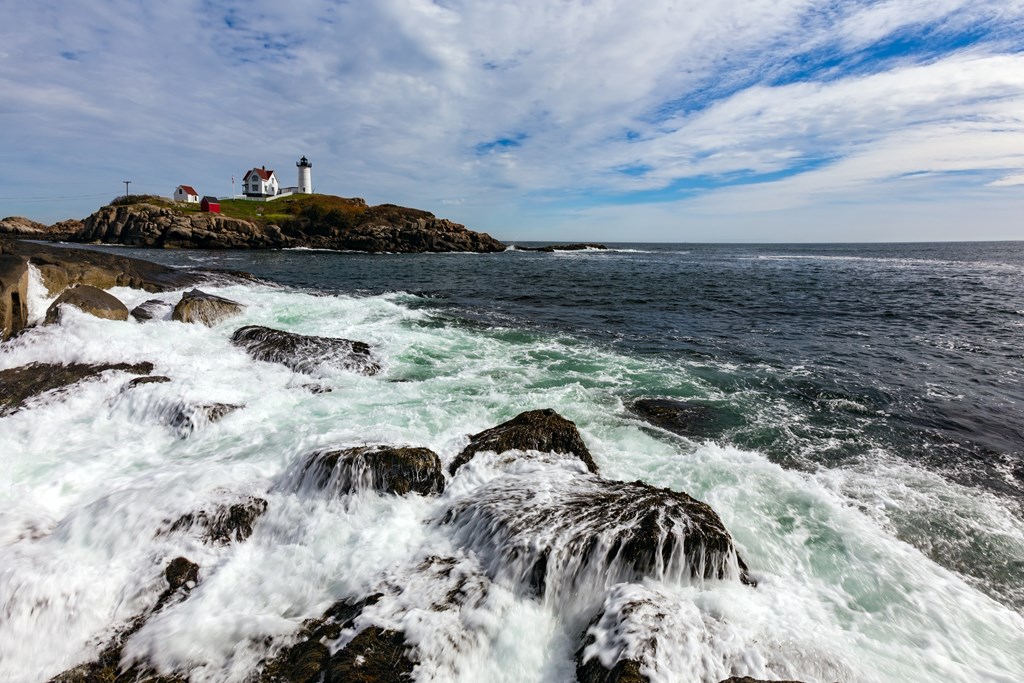 The Nubble Lighthouse stands on an island in the distance as the ocean crashes over beachside rocks.