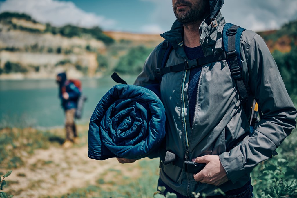 Young man on a camping trip holding sleeping bag near a mountain lake.