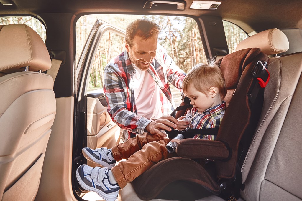 Father is adjusting child's car seat, while kid is playing with toy binoculars. Side view