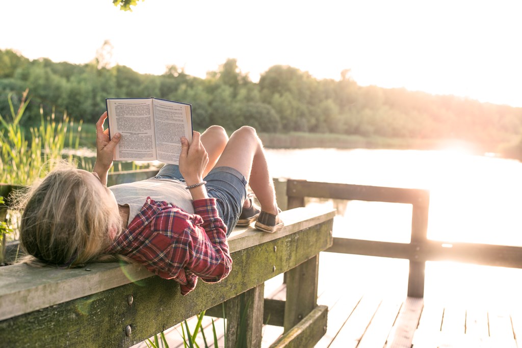 Young girl reading a book while lying on her back on a wooden beam at a wooden dock at the lake.