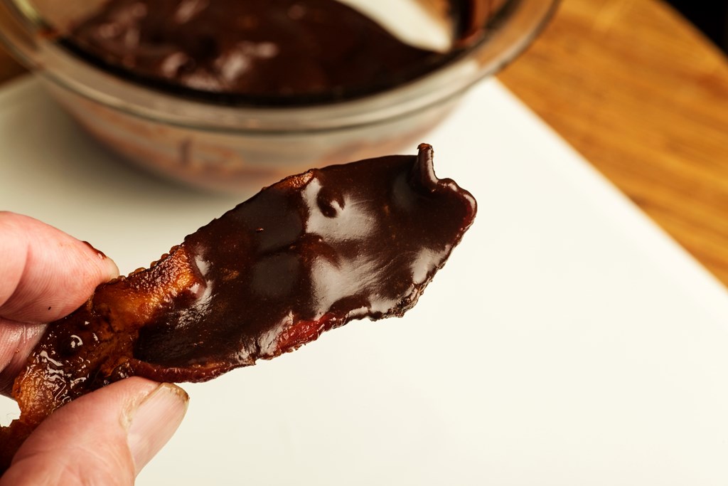 Strip of crispy bacon covered with chocolate ganache with glass bowl in the background