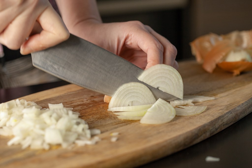 Closeup of a person cutting white onion on a wooden cutting board.