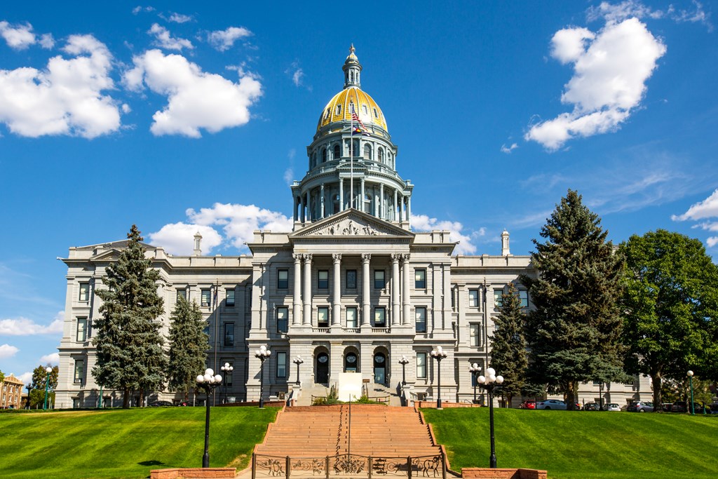 The Colorado State Capitol located in Denver. A large building with columns and large gold dome at the center against a blue sky with fluffy clouds.