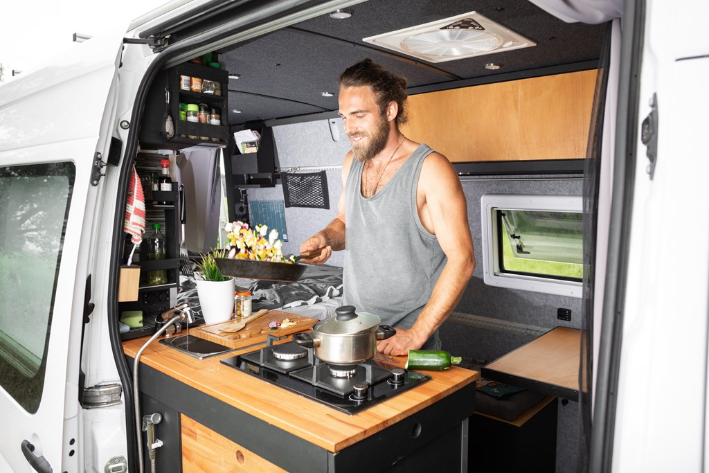 A young man flipping food in a pan in a camper van.