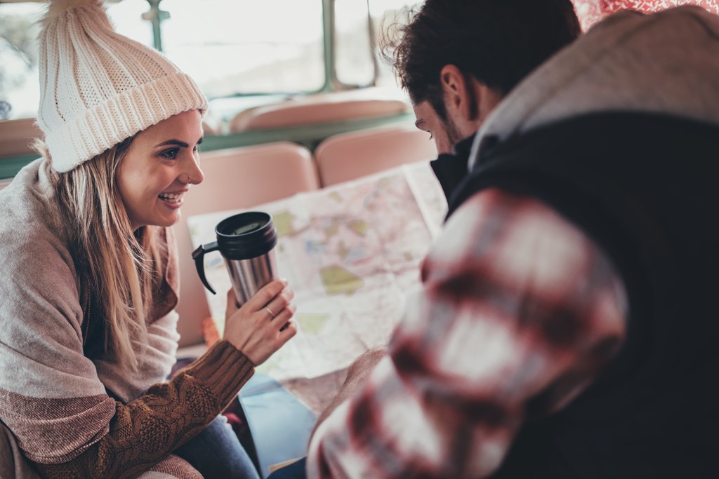 Couple of friends on roadtrip sitting at the back in their camper van. Smiling woman holding coffee and man looking at the map.