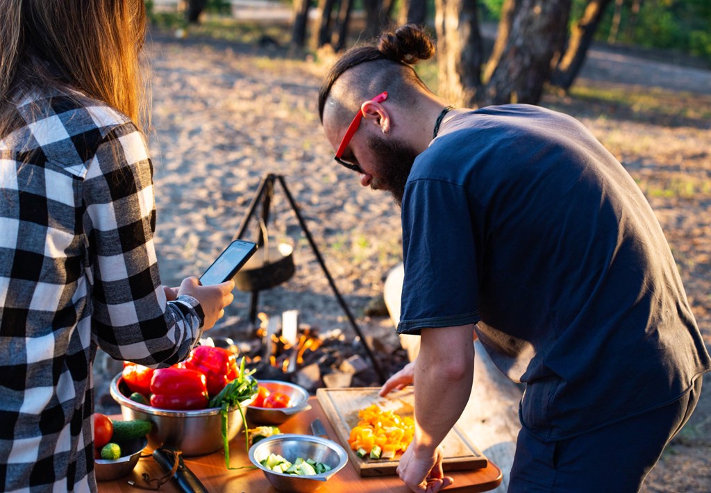 Stylish man with a beard and black glasses cuts fresh vegetables peppers and tomatoes to prepare a salad at a KOA campground.