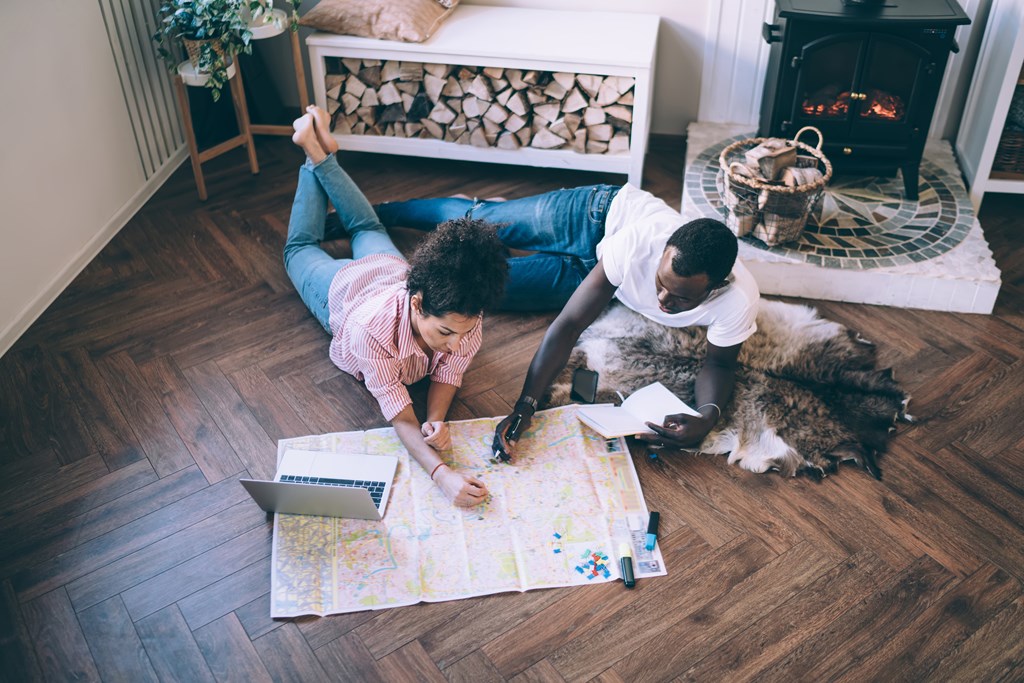 A couple has a map sprawled out on the wood floor of their home planning a trip.