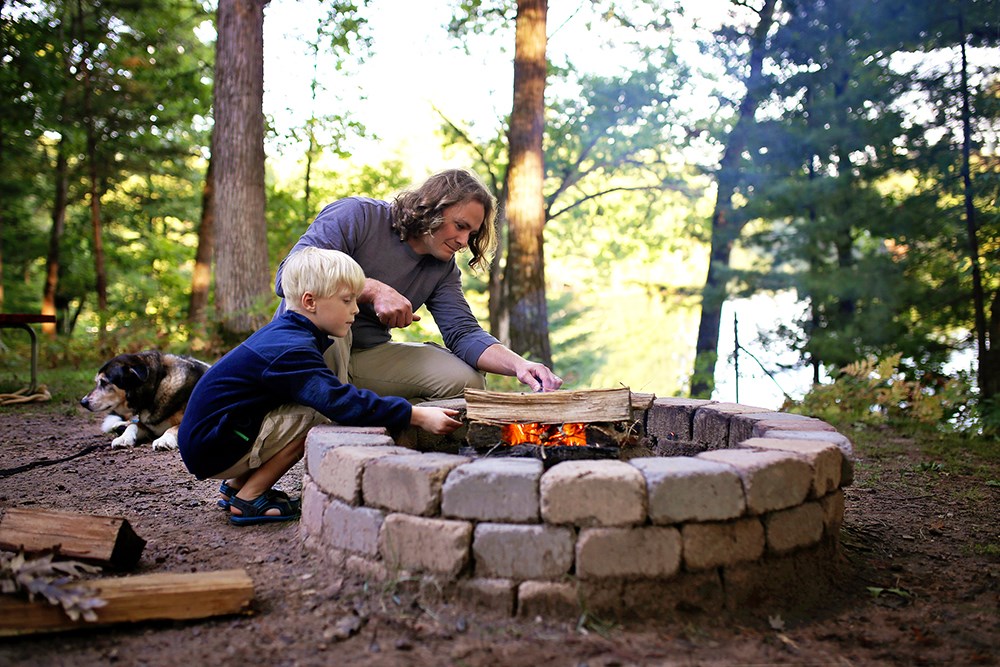 A father and his little son are starting a campfire in a fire ring at a campground overlooking a lake in the woods, as their senior dog lays nearby.