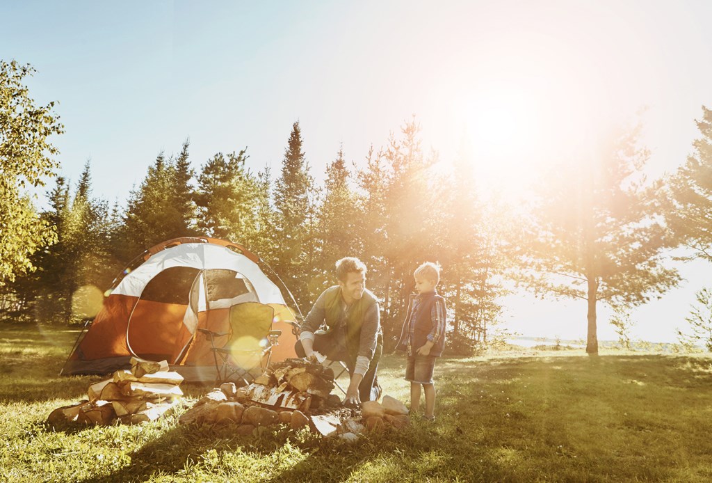 A father and son tend to the campfire near their tent on a sunny morning at a KOA campground.