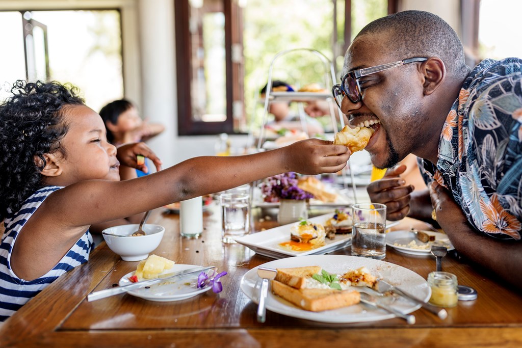 Child feeds dad a pastry at breakfast.