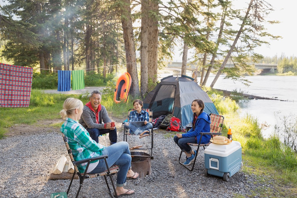 Family of four laughs around the campfire during an afternoon at a riverside campsite.