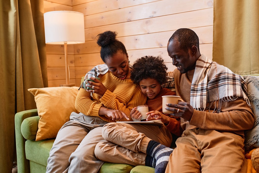 Affectionate family with hot drinks discussing something in digital tablet while relaxing on comfortable couch in a cabin.