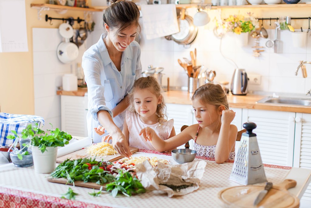 Mother making pizzas with two younger daughters in a bright kitchen.