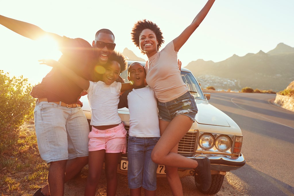 Family with two young children strike a fun pose in front of their car on a road trip.