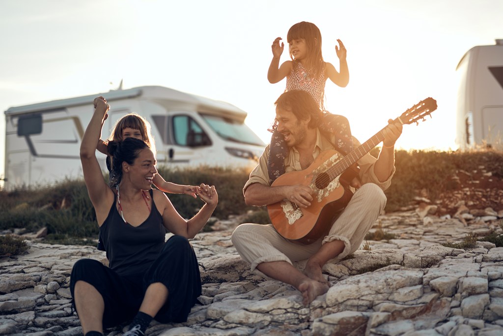 Family on a vacation, singing, playing music on a guitar and enjoying summertime vibes on a rocky beach in front of their RV.
