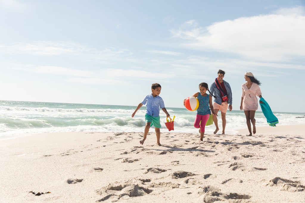 Multiracial parents walking behind children running on sand at beach during sunny day.