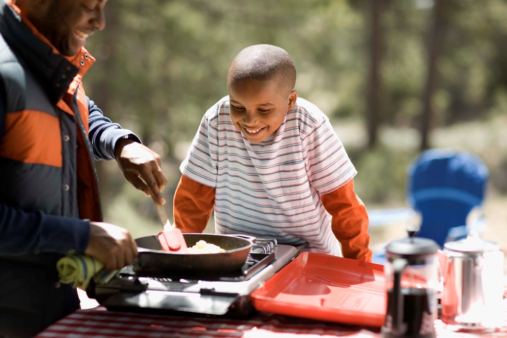 Young son looks on as father cooks scrambled eggs on a camping stove.