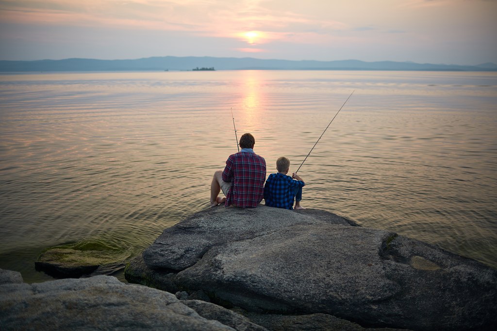 A father and son fish while sitting on stone by lake in the evening.