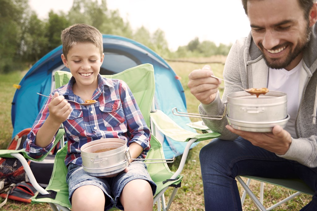 Father and son sit by a tent eating soup during a camping trip.